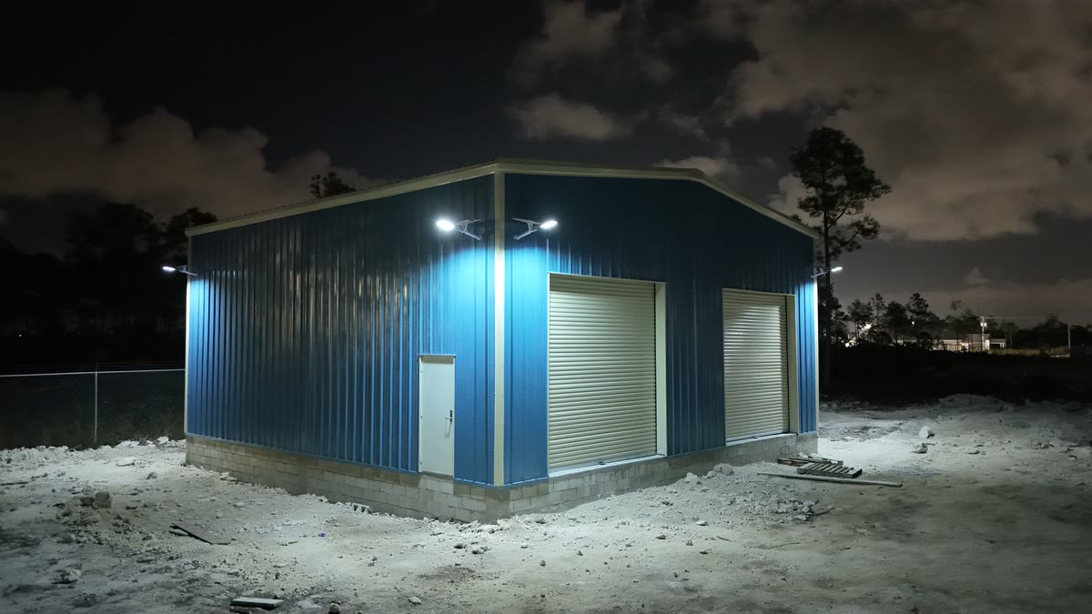 Blue steel warehouse with roll-up doors lit at night by wall-mounted solar security lights installed by JTECH